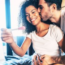 A smiling woman holds a positive pregnancy test while her partner embraces her. They look joyful in a cozy room, celebrating after managing stress with the Personalized 90-Day Fertility Program on their fertility journey.