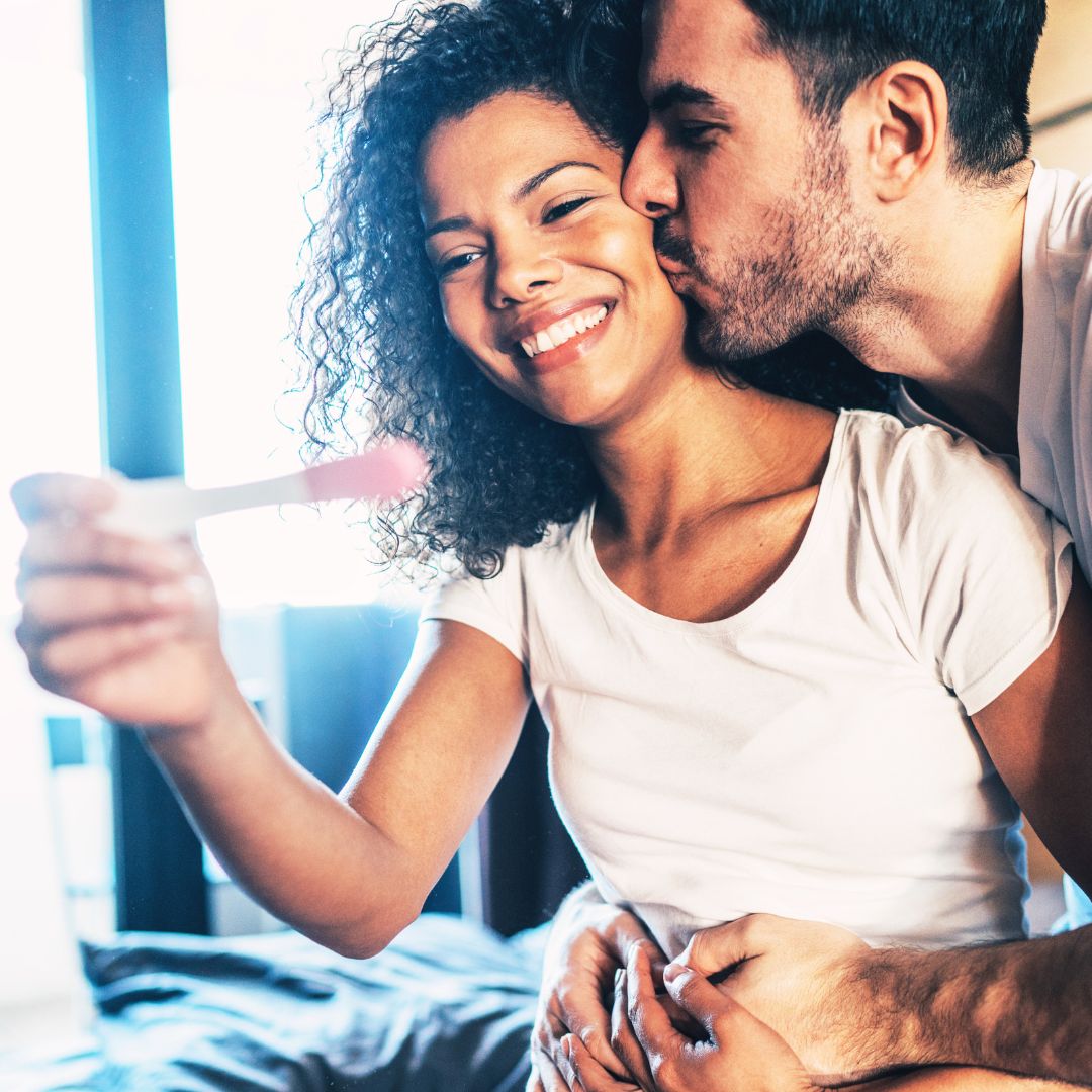 A smiling woman holds a positive pregnancy test while her partner embraces her. They look joyful in a cozy room, celebrating after managing stress with the Personalized 90-Day Fertility Program on their fertility journey.