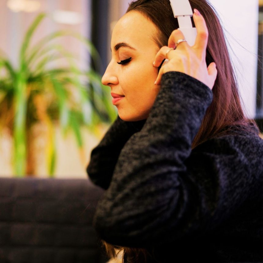 A woman with long brown hair, in a dark sweater, relaxes with her eyes closed, listening to You’re doing everything but nothing’s work on white headphones while a blurred plant sits in the background.
