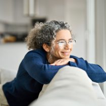 Woman with gray curly hair and glasses smiling thoughtfully beside a window.