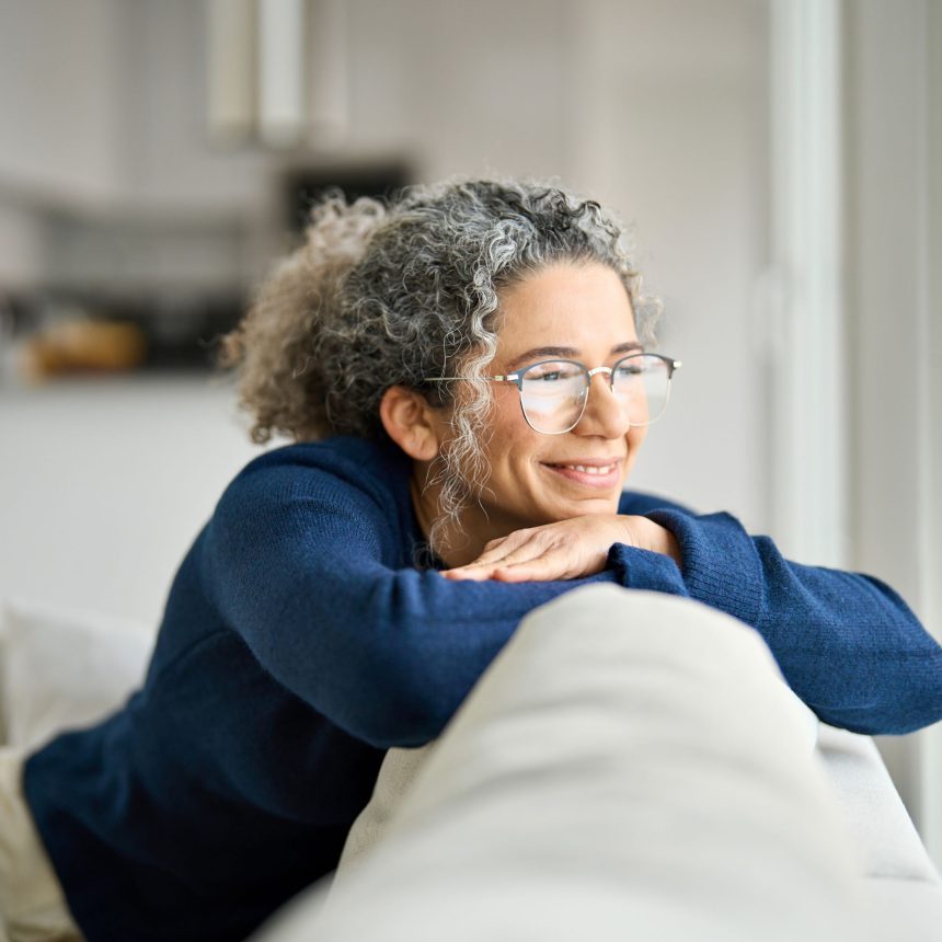 Woman with gray curly hair and glasses smiling thoughtfully beside a window.