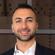 A smiling man with short dark hair and trimmed beard, wearing a white shirt and dark blazer, stands in front of a background with metal studs and panels.