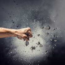 A human fist punches through a cloud of shattered black debris and dust, symbolizing the power of self-expression in Find Your Power, with particles flying outward against a dark, smoky background.