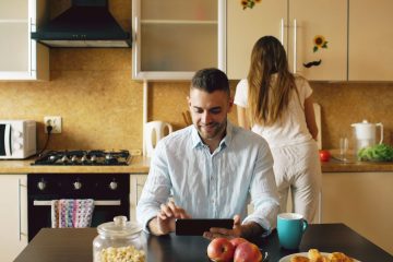 A man sits at a kitchen table using a tablet, smiling. There are apples, popcorn, and a coffee mug on the table. In the background, his wife stands at the counter with her back turned, preparing food—perhaps an example of weaponized incompetence in relationships as he appears content to leave all the kitchen work to her.