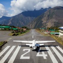 A small airplane readies for takeoff on runway 24 at a Nepal mountain airport, gateway to the Everest Base Camp Trek 2026, surrounded by green buildings and forested hills beneath a partly cloudy sky.