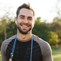 A smiling, bearded man stands outdoors wearing Release Your Pain wireless earbuds, a black athletic shirt, and a jump rope around his neck, with green trees and sunlight in the background.