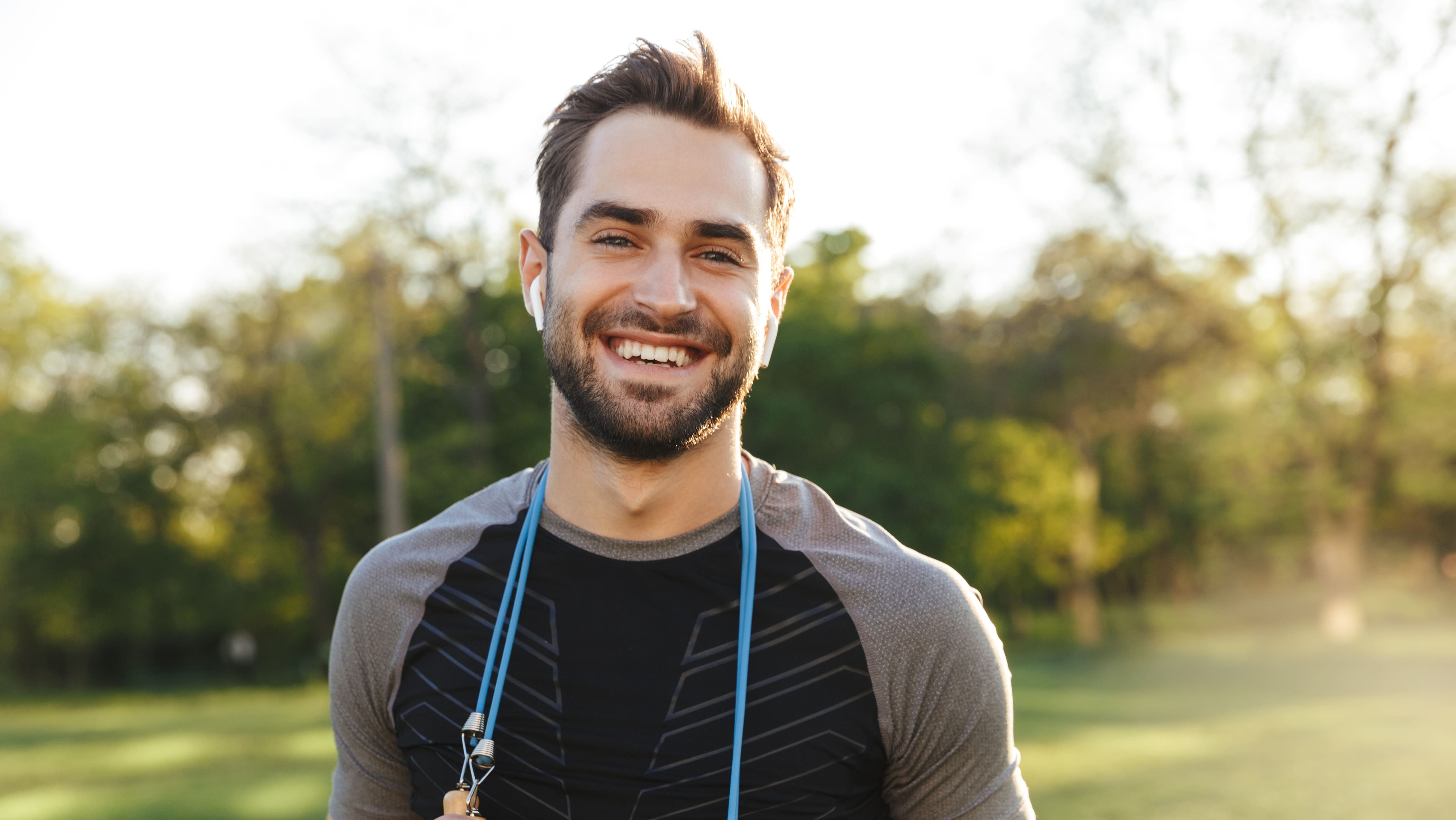 A smiling, bearded man stands outdoors wearing Release Your Pain wireless earbuds, a black athletic shirt, and a jump rope around his neck, with green trees and sunlight in the background.