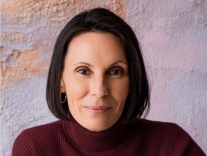 A woman with straight dark hair, wearing a maroon turtleneck and hoop earrings, smiles gently before a textured light wall, reflecting the harmony and balance achieved through Kinesiology Deep Healing & Transformation.