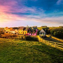 Colorful houses line a rural road at sunset, with bright pink, yellow, and orange facades. A river flows nearby, and rolling green fields stretch into the distance under a vivid, dramatic sky.