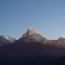 Experience the Annapurna Base Camp Trek Nepal as snow-capped peaks glow in golden sunrise light against a clear blue sky, creating breathtaking views above shadowy foothills—an unforgettable Himalayan adventure.