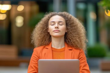 Woman with curly hair in an orange blazer sits outdoors, eyes closed in peaceful meditation and work balance, holding a laptop. She appears relaxed, highlighting the benefits of meditation amid greenery and soft lights.