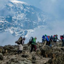 A group of hikers with backpacks trek over rocky terrain toward a snow-capped mountain, embodying the spirit of adventure on the Kilimanjaro Expedition.