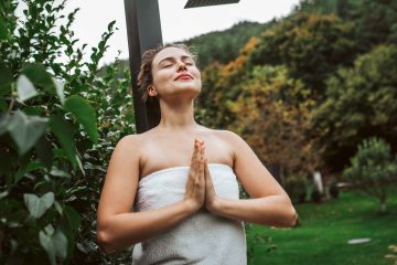 A woman wrapped in a white towel stands outdoors, hands in prayer position and eyes closed, enjoying peace. Surrounded by lush greenery, she appears to be engaging in daily readings and meditations in a tranquil natural setting.