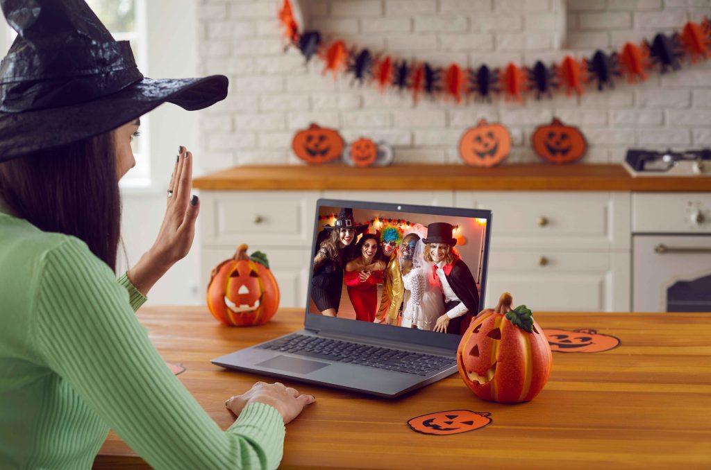 Woman wearing a witch hat waving hello at her team, which appear on a screen on her laptop. Two pumpkins sit next to the laptop on the kitchen table.