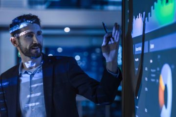 A man in a suit confidently points at a large digital screen displaying colorful graphs and charts, appearing to show how to not get nervous when presenting in a modern, dimly lit office setting.