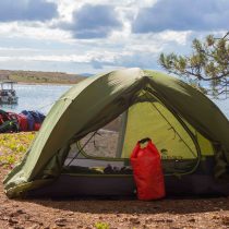 A green tent and red dry bag sit on a grassy lakeside campsite, perfect for a Croatia – Sea Kayak Adventure. Kayaks, a dock, and people near the water appear in the background beneath a partly cloudy sky.