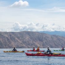Five people paddle colorful kayaks on calm water by rocky cliffs under a cloudy sky, enjoying the Croatia – Sea Kayak Adventure.