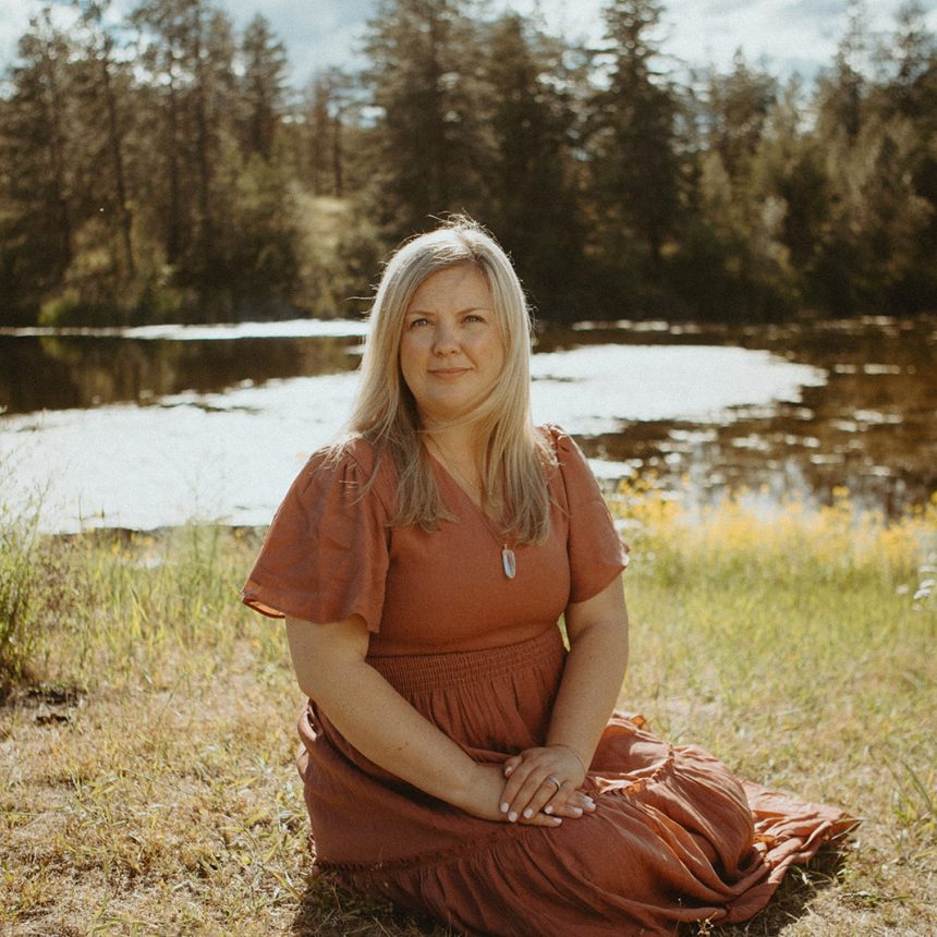 A woman with long blonde hair in a rust-colored dress sits on the grass by a pond, surrounded by wildflowers and trees, under a partly cloudy sky.