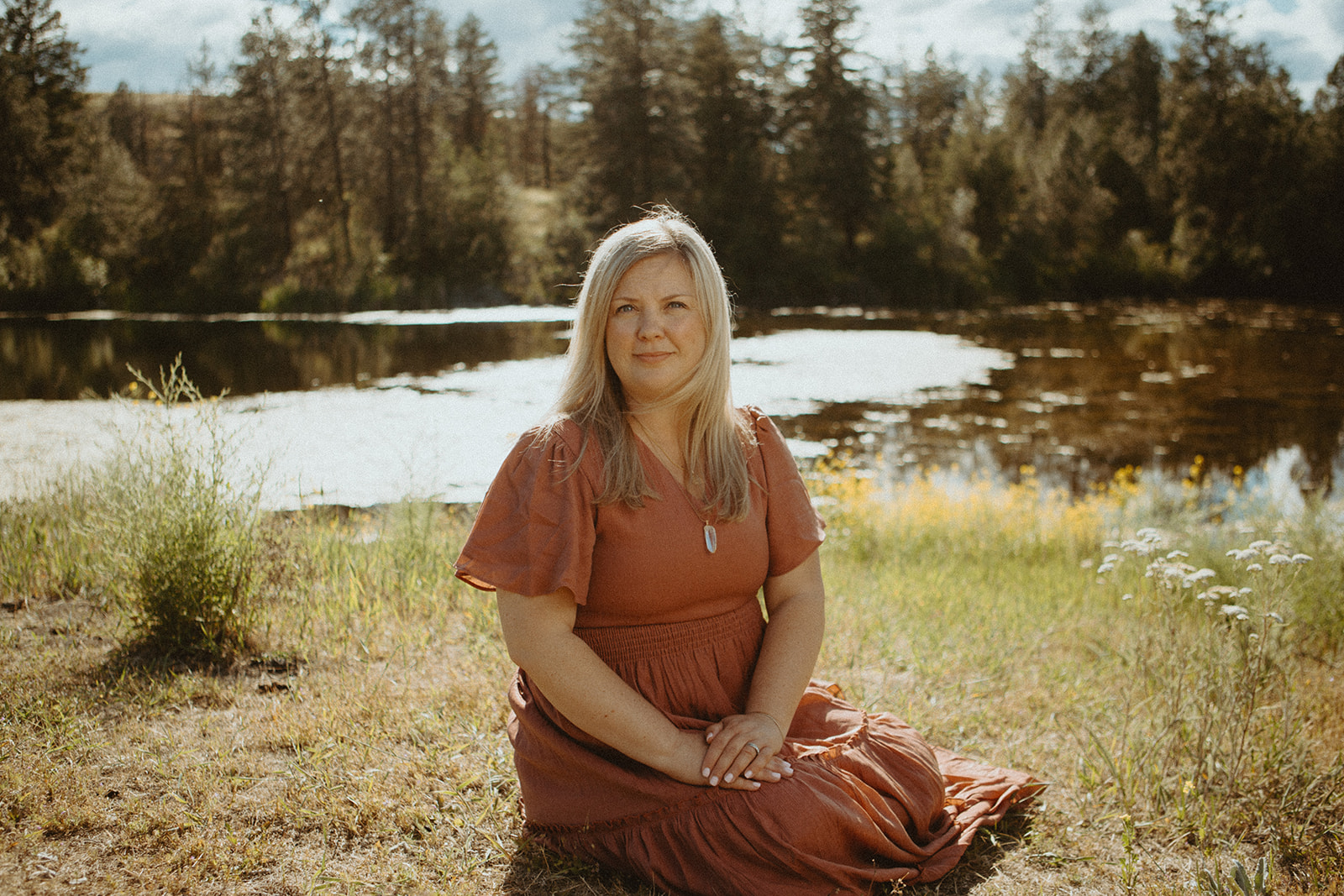 A woman with long blonde hair in a rust-colored dress sits on the grass by a pond, surrounded by wildflowers and trees, under a partly cloudy sky.