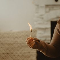 A person wearing a brown long-sleeve shirt holds a lit matchstick indoors, near a white brick fireplace and a textured beige surface in the background.