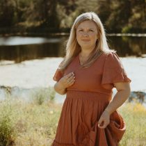 A woman with long blonde hair wearing a rust-colored dress stands outdoors near a pond, surrounded by greenery and wildflowers, with sunlight filtering through the trees.