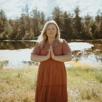 A woman in a rust-colored dress stands outdoors near a pond, eyes closed, with hands pressed together in a prayer gesture. Sunlight filters through trees in the background, creating a peaceful, serene atmosphere.