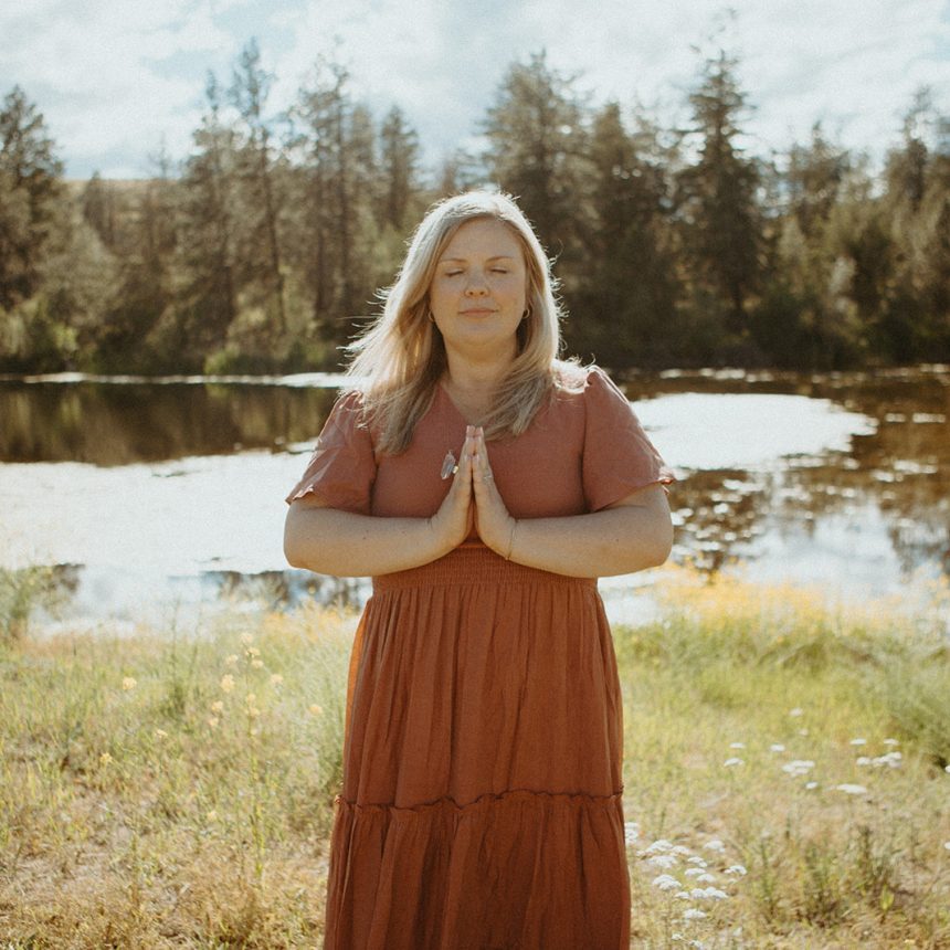 A woman in a rust-colored dress stands outdoors near a pond, eyes closed, with hands pressed together in a prayer gesture. Sunlight filters through trees in the background, creating a peaceful, serene atmosphere.