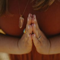A person wearing a brown top and a crystal pendant necklace holds their hands together in a prayer or namaste gesture, showing white-tipped nails and rings on their fingers.