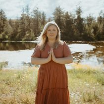 Photo of Jennifer Kanyumwa the owner of Mujen Community. She is standing beside a pond with trees in the background. She has blonde hair and is wearing a burnt orange dress with her hands in prayer.