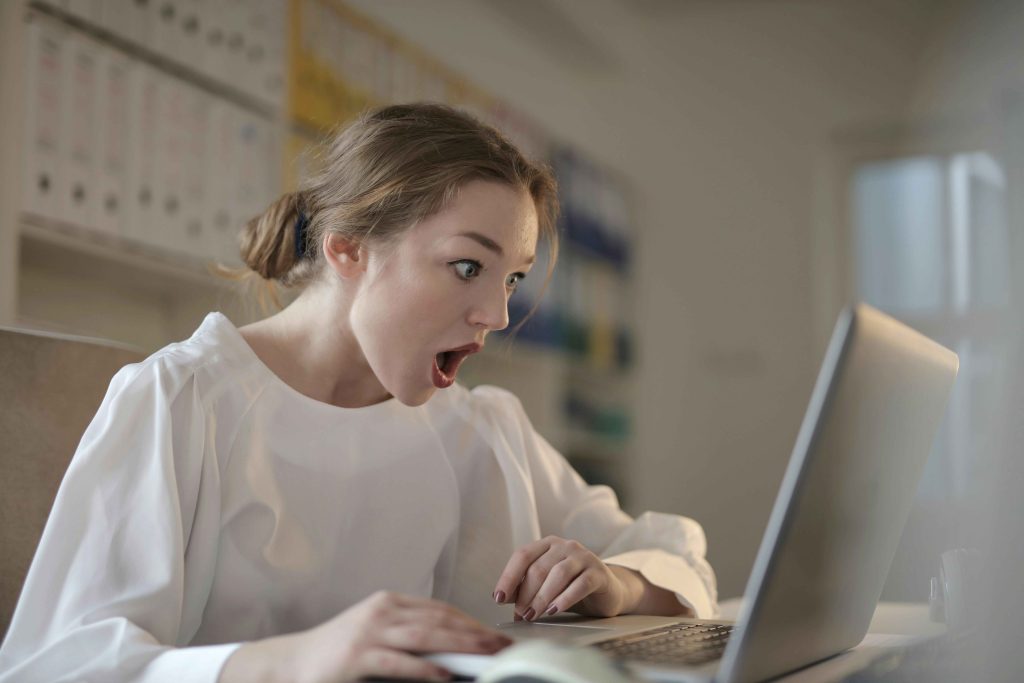 A woman in a white blouse with a highly expressive face looking at her laptop.