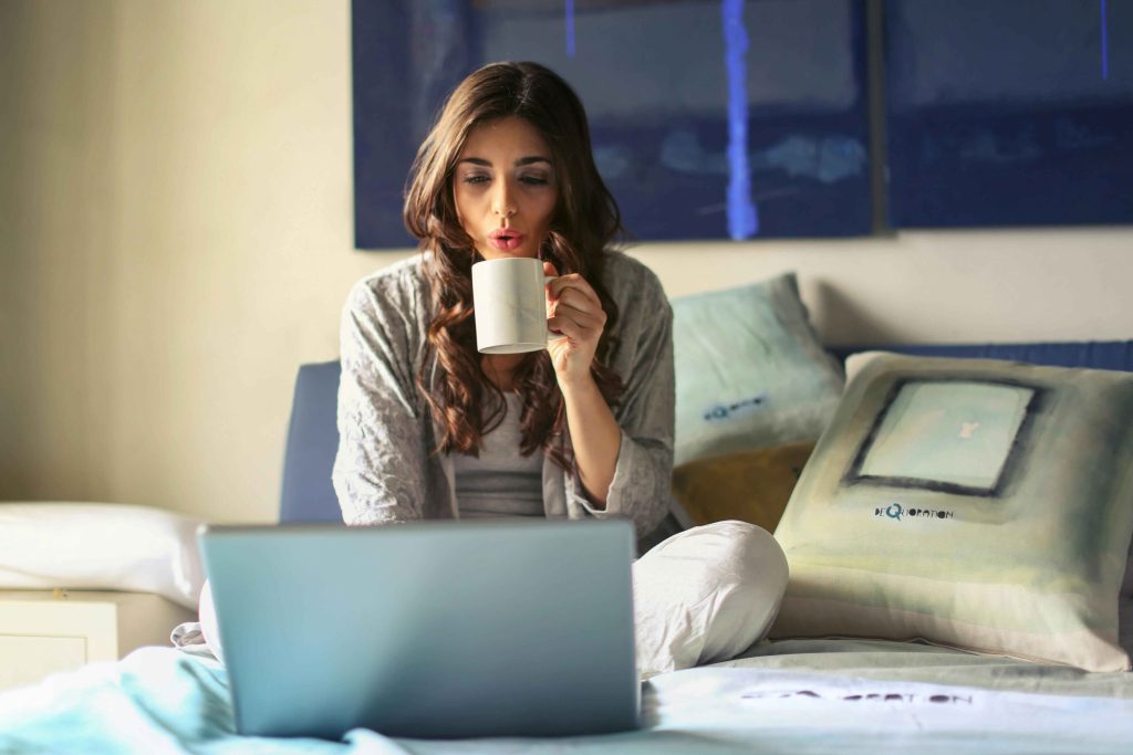 Young woman sitting on her bed drinking a cup of coffee while working on her open laptop
