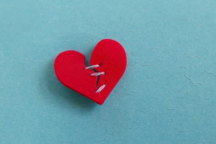A red paper heart with stitches down the middle lies on a light blue textured surface, symbolizing a mended or healing heart.