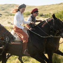 Two people ride side by side through grassy fields, embodying the spirit of the Cowgirl Yoga™ - Argentina retreat. Sunlight illuminates the rural landscape as they explore in vibrant outfits and matching berets.