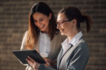 Two women in business attire are smiling while looking at a tablet, discussing the benefits of career coaching. One holds a coffee cup as they stand in front of a blurred brick wall background.