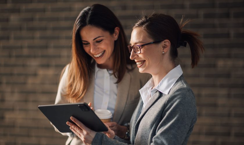 Two women in business attire are smiling while looking at a tablet, discussing the benefits of career coaching. One holds a coffee cup as they stand in front of a blurred brick wall background.
