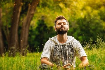 A man sits cross-legged in tall grass in a sunlit forest, eyes closed and smiling peacefully, surrounded by green trees and floating pollen or dust particles in the air.
