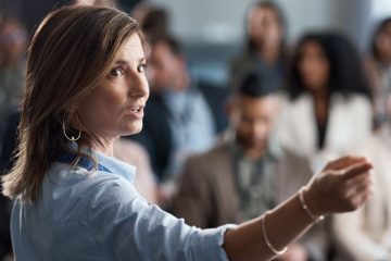 Woman speaking to a group with her hand raised as others listen