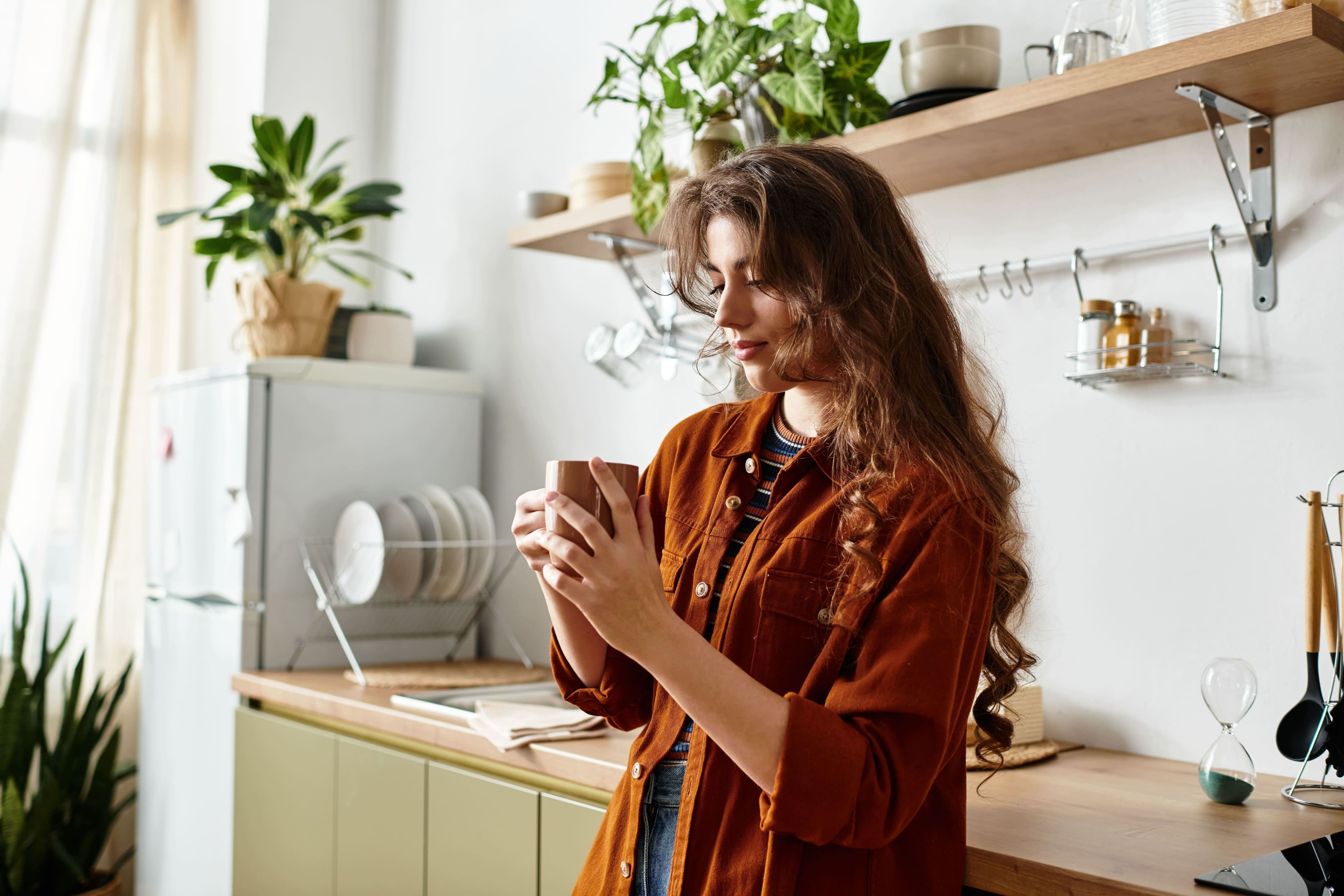 A woman with long brown hair stands in a bright kitchen, holding a mug and smiling—her confident posture subtly reflecting the power of assertiveness. There are plants, shelves with dishes, and sunlight streaming through a window in the background.