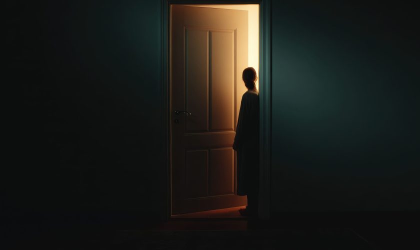A silhouette of a person stands in a doorway, illuminated by warm light from the other side, while the surrounding room is dark and shadowy.