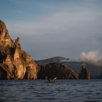 A person paddles on the Sicily Sea Kayak Expedition, gliding over calm blue water bordered by steep rocky cliffs and distant volcanic mountains beneath a partly cloudy sunset sky.