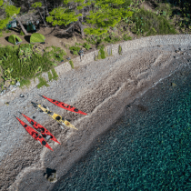 Aerial view of three kayaks—two red, one yellow—on a rocky Sicily shoreline by clear blue water. Two green tents from the Sicily Sea Kayak Expedition are set up under trees behind a stone wall on the shore.