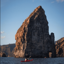 A person paddles a red kayak on calm water near jagged rocks at sunset, with rugged cliffs behind—an idyllic scene from the Sicily Sea Kayak Expedition under a clear blue sky.