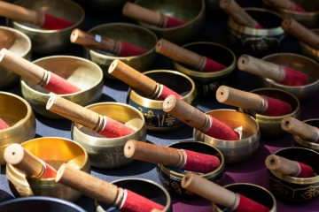 A collection of Tibetan singing bowls with wooden mallets, arranged closely together. The bowls are gold and black with decorative markings, and sunlight highlights their metallic surfaces.