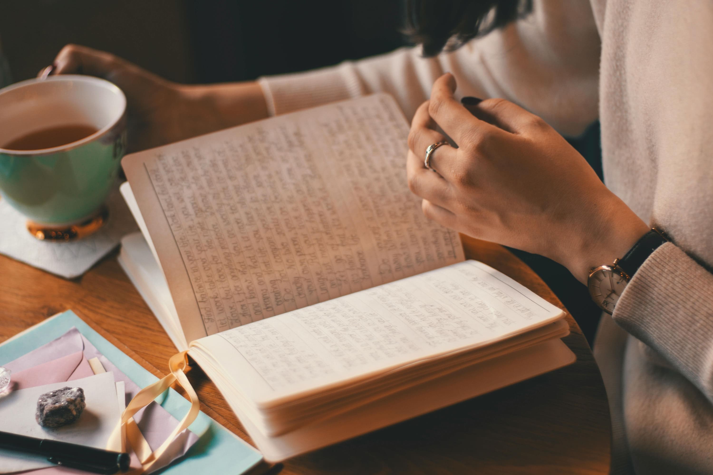 A person holding a cup of tea sits at a wooden table, writing in an open journal filled with handwritten notes. Colored envelopes, stones, and a ribbon bookmark are nearby. The person wears a watch and a ring.