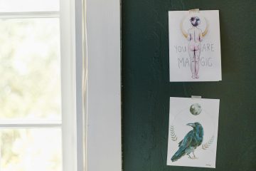 Two illustrated cards taped to a dark green wall; the top card features a mystical figure and the words YOU ARE MAGIC, while the bottom card depicts a blue raven with a moon above its head.