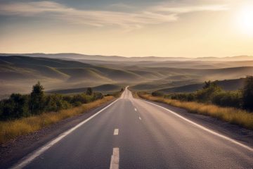 A straight, empty road stretches into the distance through rolling green hills under a clear sky at sunset, with soft sunlight illuminating the landscape and distant mountains.