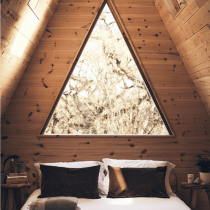 Enjoy three nights of spring yoga in a cozy wooden A-frame cabin bedroom, featuring a large triangular window behind the bed, white linens, brown pillows, and a folded blanket at the foot. Rest and Re-wild awaits you.