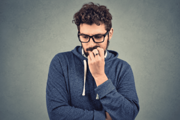 A man with curly hair and glasses wearing a blue hoodie looks down thoughtfully, with his hand touching his mouth in a gesture that suggests he is worried or deep in thought, perhaps contemplating conscious decision making and how to make better decisions.