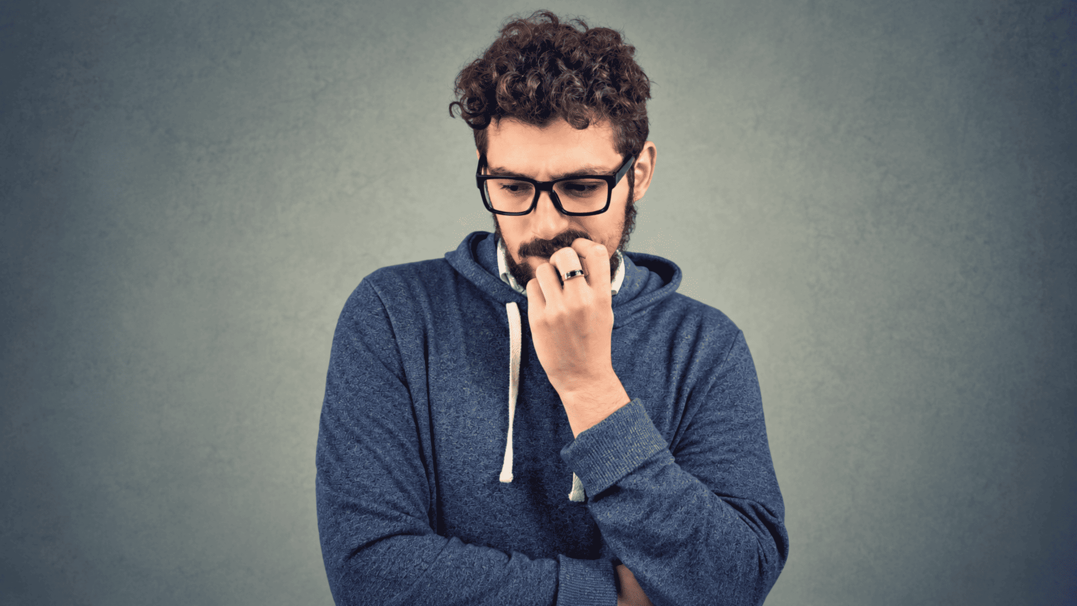 A man with curly hair and glasses wearing a blue hoodie looks down thoughtfully, with his hand touching his mouth in a gesture that suggests he is worried or deep in thought, perhaps contemplating conscious decision making and how to make better decisions.