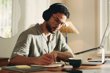 Man wearing headphones writes in a notebook beside his laptop